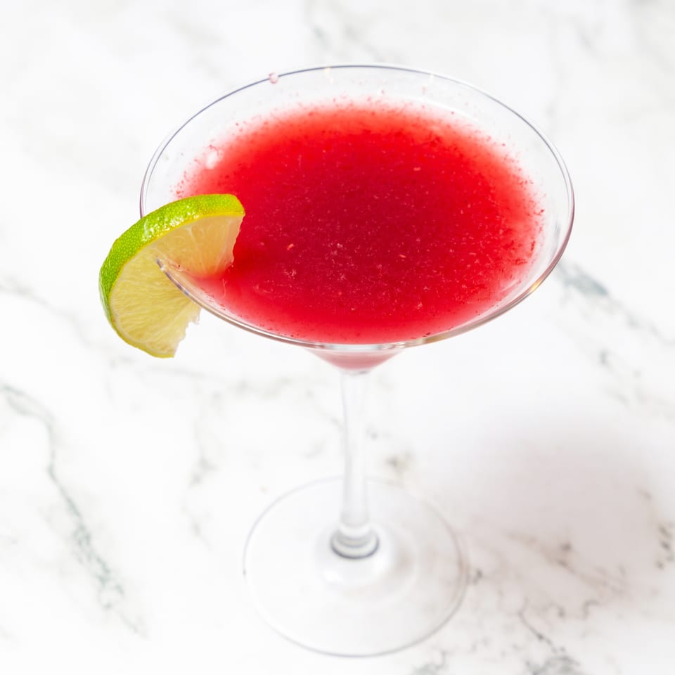 Bright red cocktail in a martini glass garnished with a lime wedge on a white marble surface.