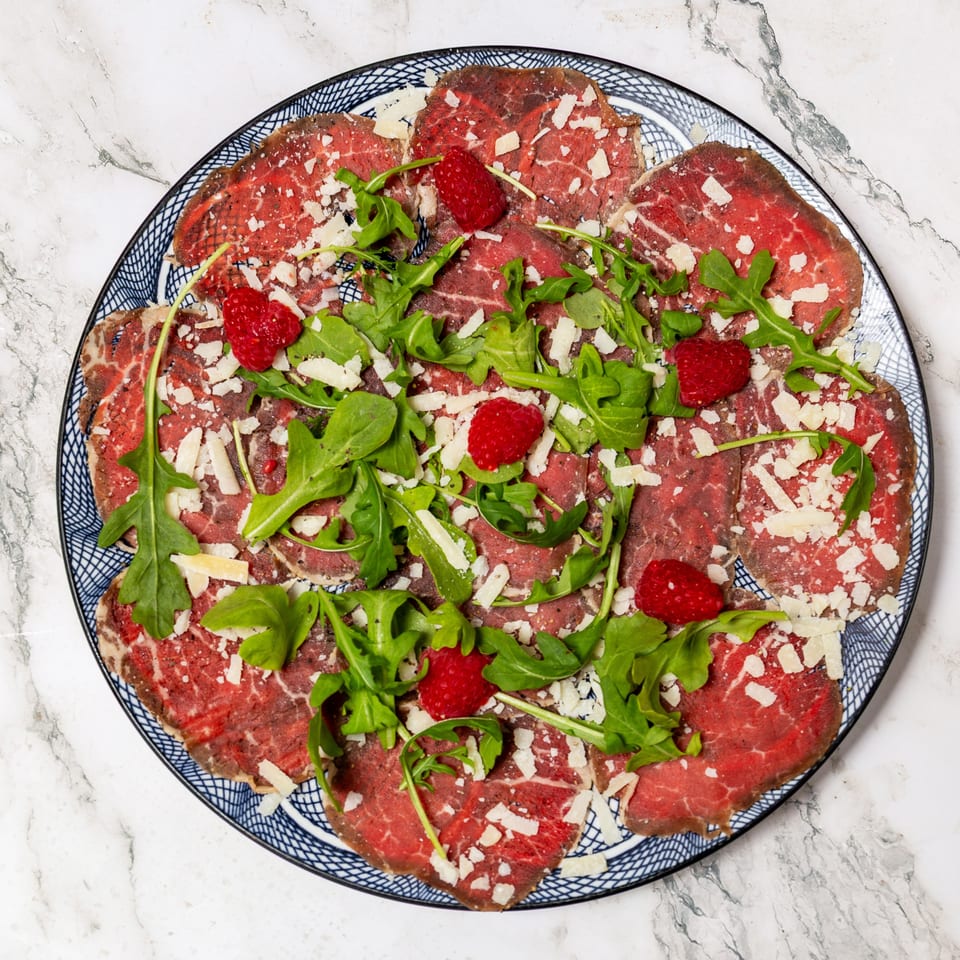 Thinly sliced beef carpaccio topped with arugula, Parmesan shavings, and raspberries on a patterned plate.
