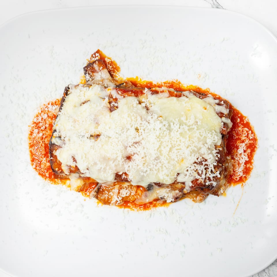 Baked eggplant Parmesan layered with tomato sauce, melted cheese, and grated Parmesan on a white plate.