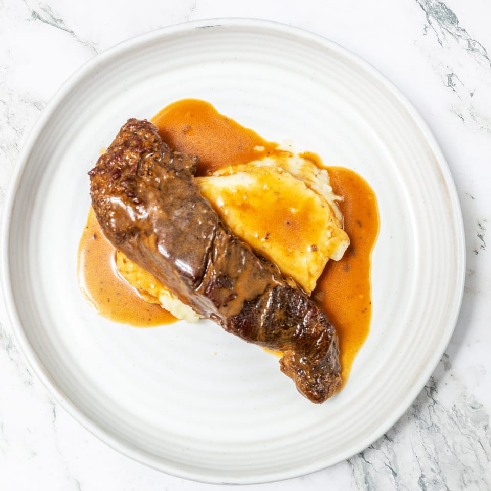 Grilled steak served over creamy mashed potatoes with brown gravy on a white plate.
