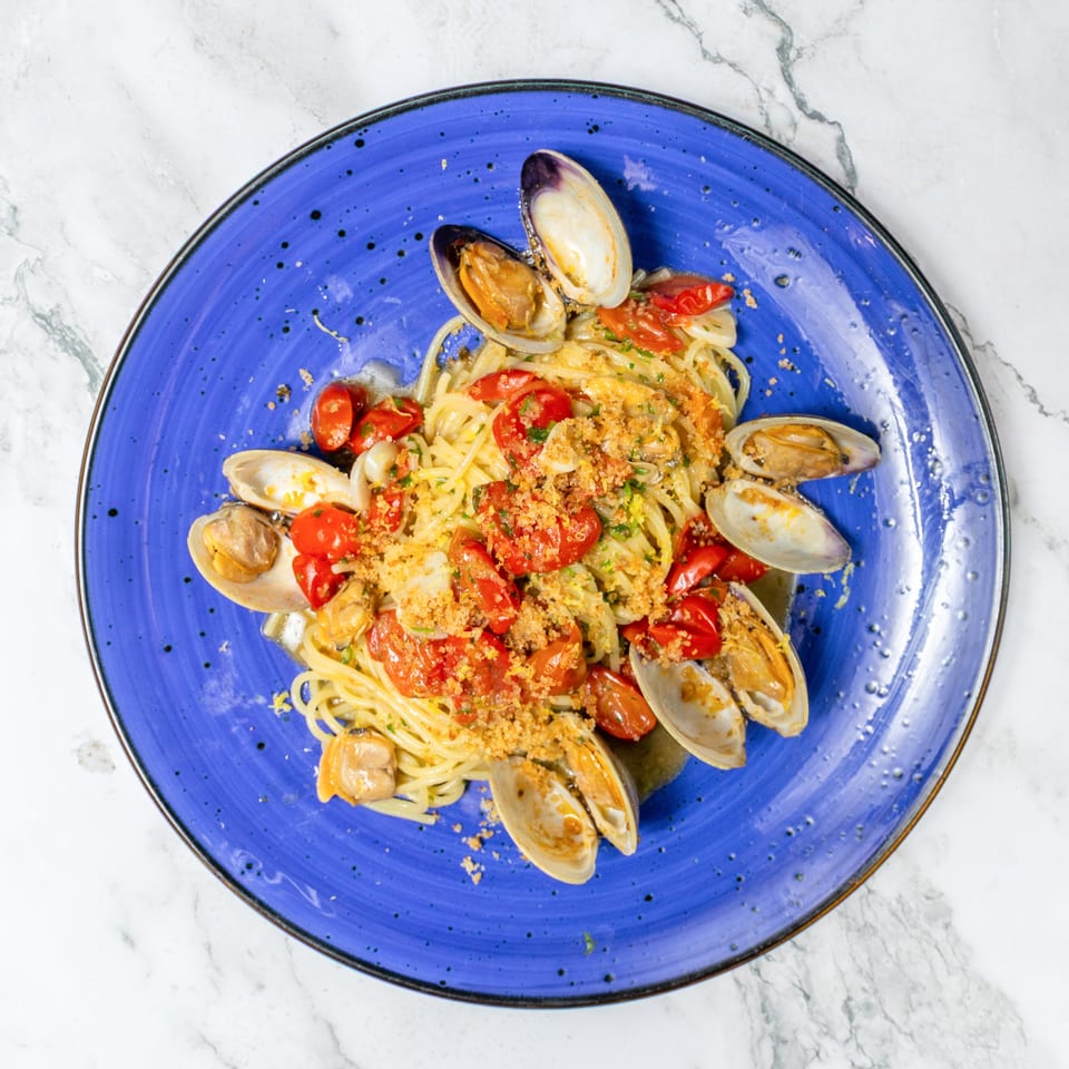 Spaghetti with clams, mussels, cherry tomatoes, herbs, and toasted breadcrumbs on a blue plate.