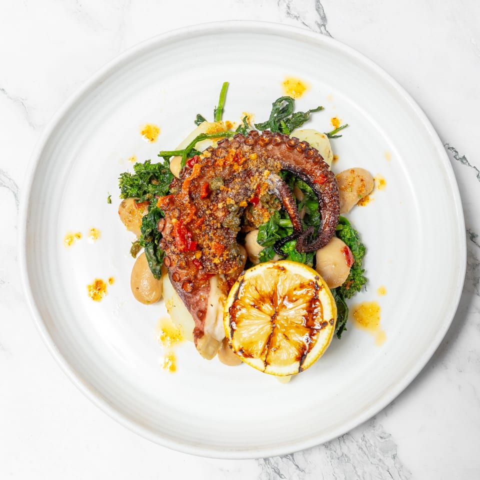 Grilled octopus with leafy greens, potatoes, spices, and a charred lemon half on a white plate.