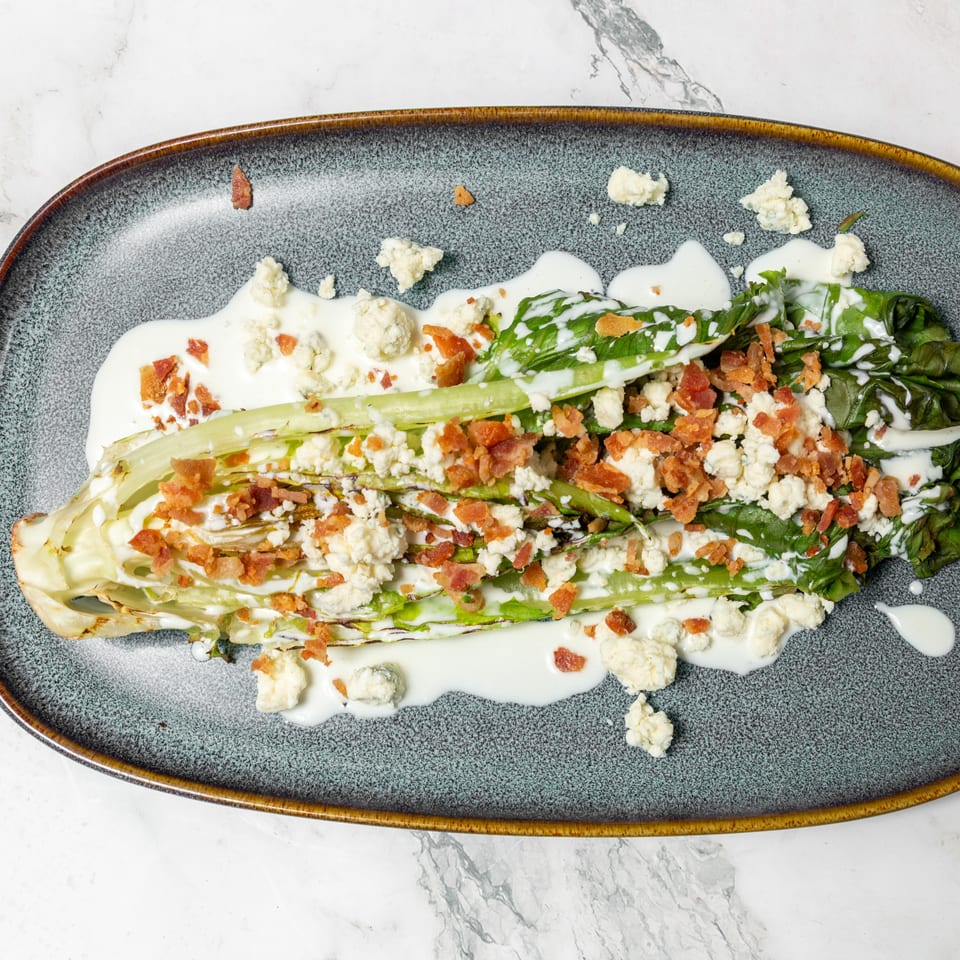 Grilled romaine lettuce topped with creamy dressing, blue cheese crumbles, and crispy bacon on a ceramic platter.