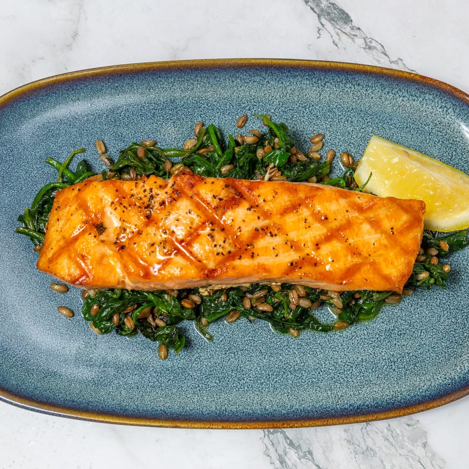 Seared salmon fillet served over sautéed greens with toasted grains and lemon wedges on a blue platter.