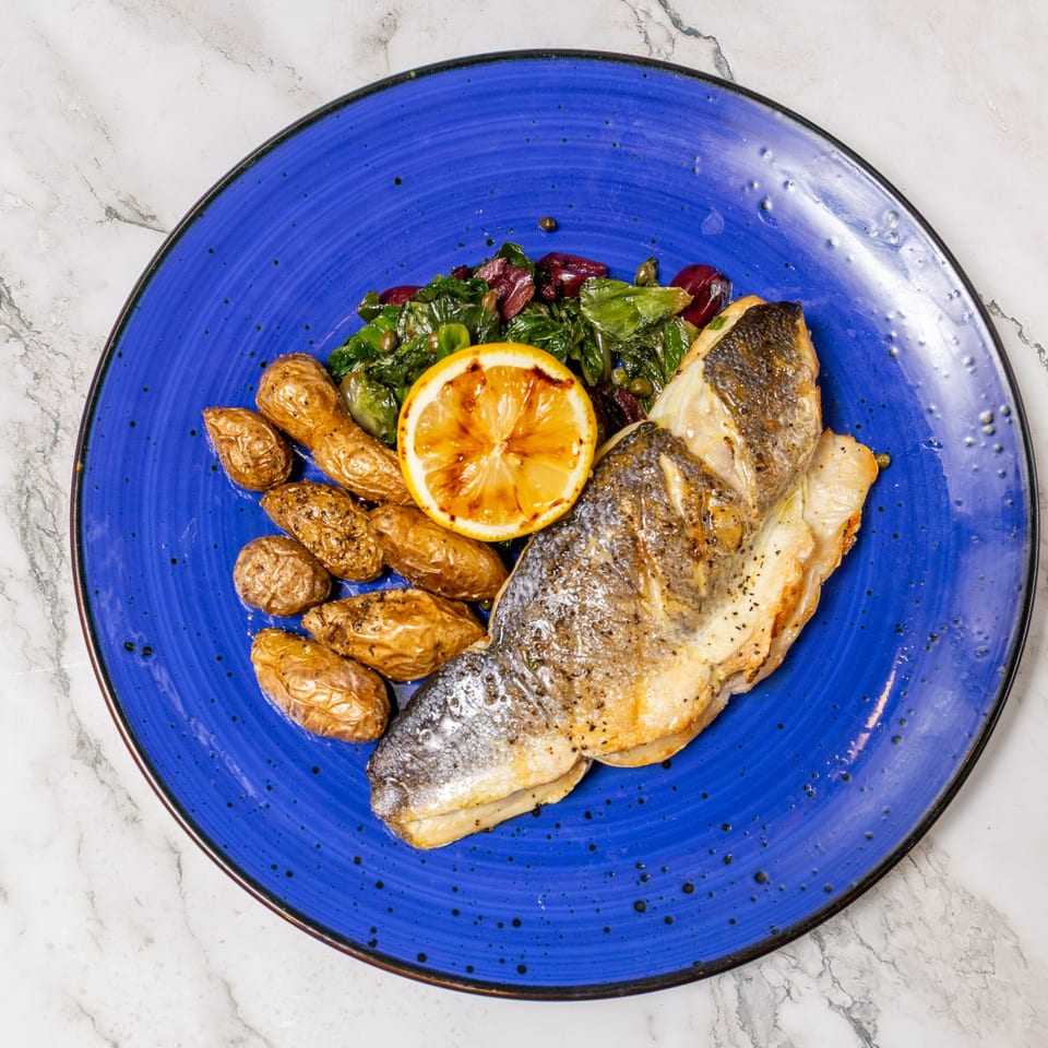 Pan-seared fish with roasted fingerling potatoes, leafy greens, olives, capers, and grilled lemon on a blue plate.