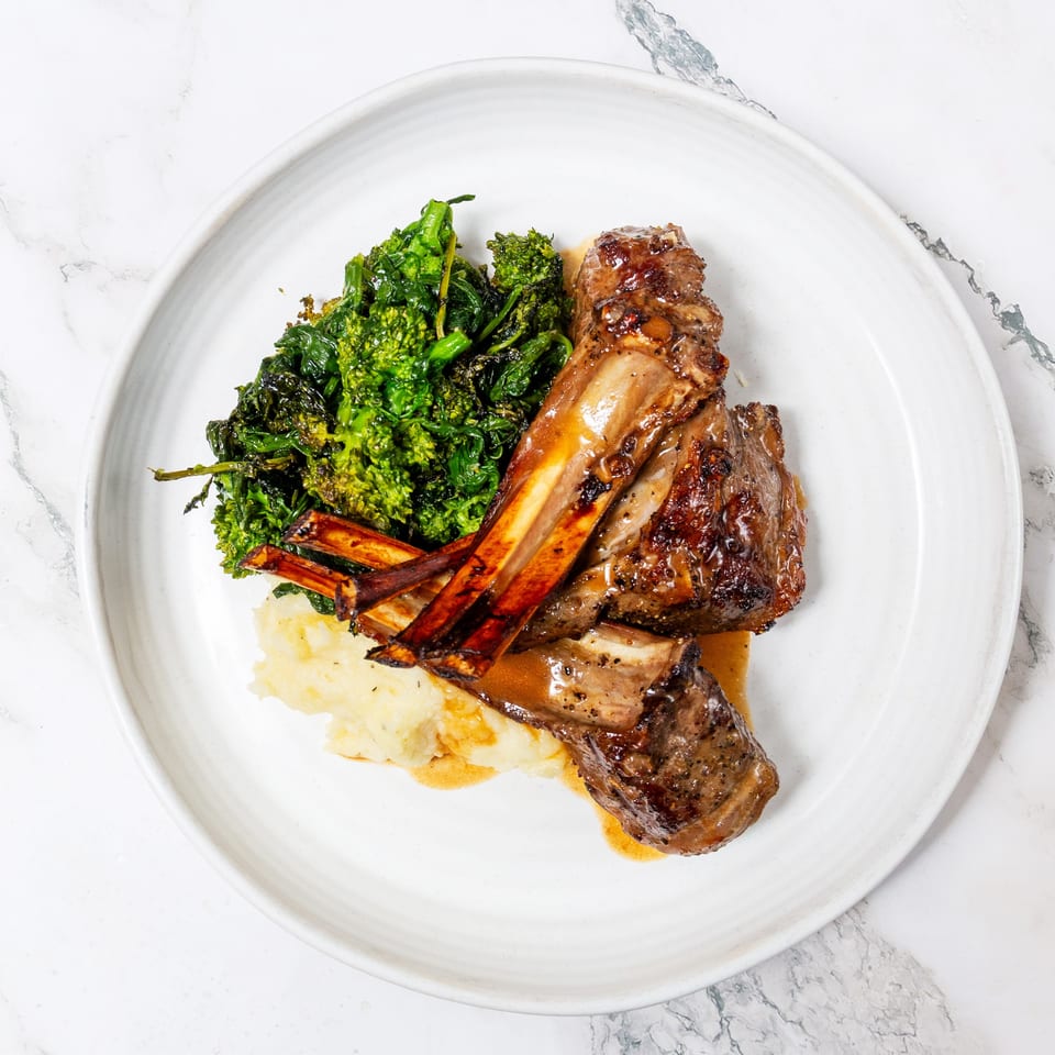 Grilled lamb chops with roasted carrots, broccolini, mashed potatoes, and pan sauce on a white plate.