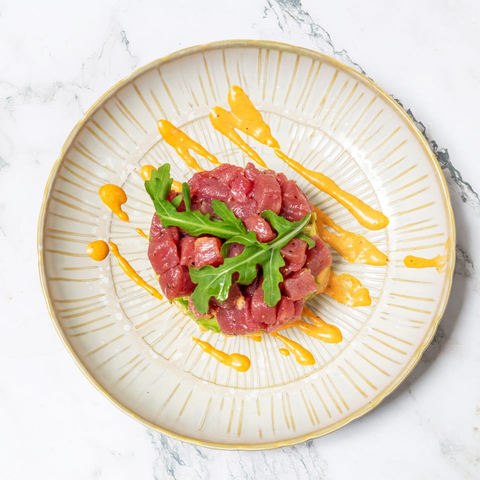 Tuna tartare topped with arugula, served on a decorative plate with orange sauce drizzles.