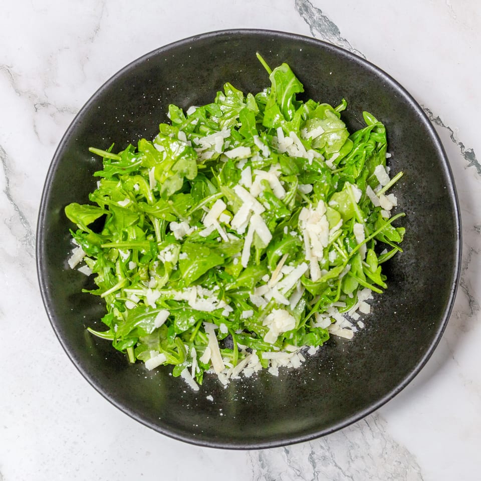 Fresh arugula salad topped with shaved Parmesan in a black bowl on a marble countertop.