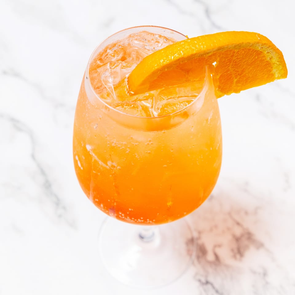 Orange cocktail with ice and a fresh orange wedge garnish in a stemmed glass on a marble surface.