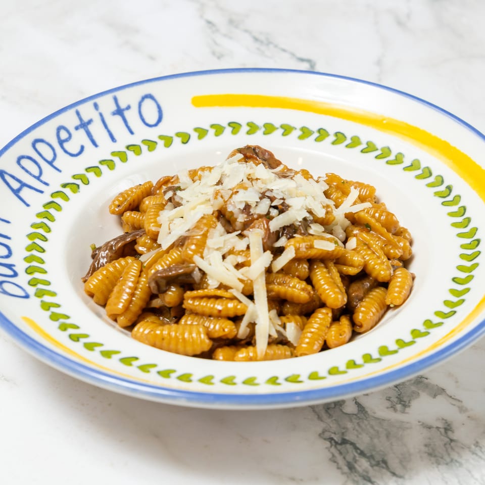 A plate of ridged pasta with mushrooms and grated cheese on a colorful Apetito dish atop a marble table.